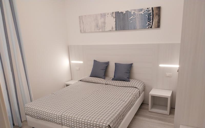 Camera moderna con letto matrimoniale, due cuscini blu e illuminazione minimalista.